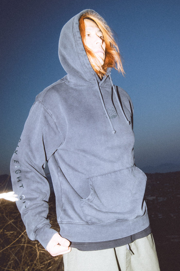 A person with long, light hair stands outdoors at dusk, looking upward while wearing the hood up on a blue 686 Men's High Def Relaxed LS Hoody. Their hair is blown across their face as the background fades into darkness with only a faint landscape visible. The cotton hoody from 686 adds comfort and warmth to the cool evening scene.