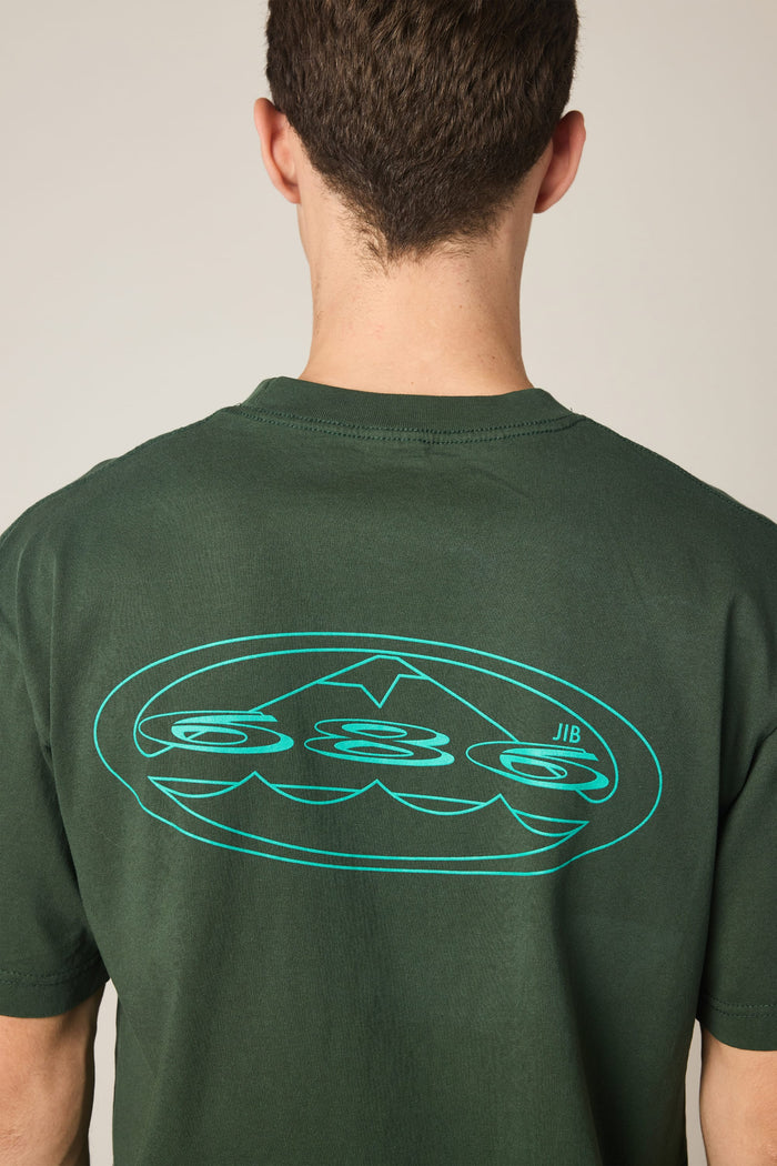 A person wearing the 686 Men's Jib Relaxed SS Tee in dark green, viewed from behind. This 100% cotton t-shirt by 686 features a large, stylized blue graphic with abstract shapes and lines on the upper back.