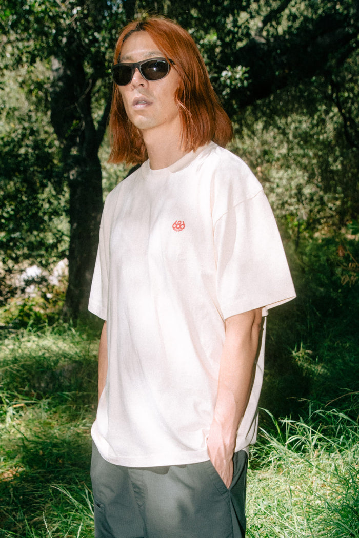 A person with shoulder-length red hair wearing sunglasses and the 686 Men's Unit Relaxed SS Tee by 686, paired with gray shorts, stands outdoors in a sunlit, wooded area with green grass and trees in the background.