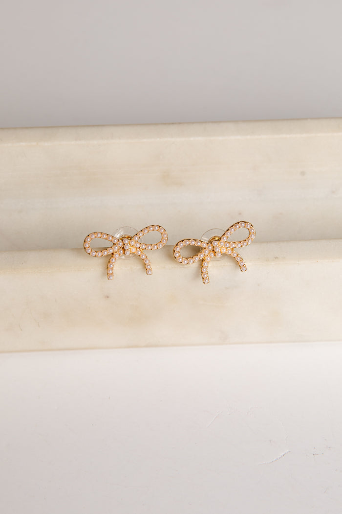 Gold bow-shaped earrings on a beige cushion with a light gray background
