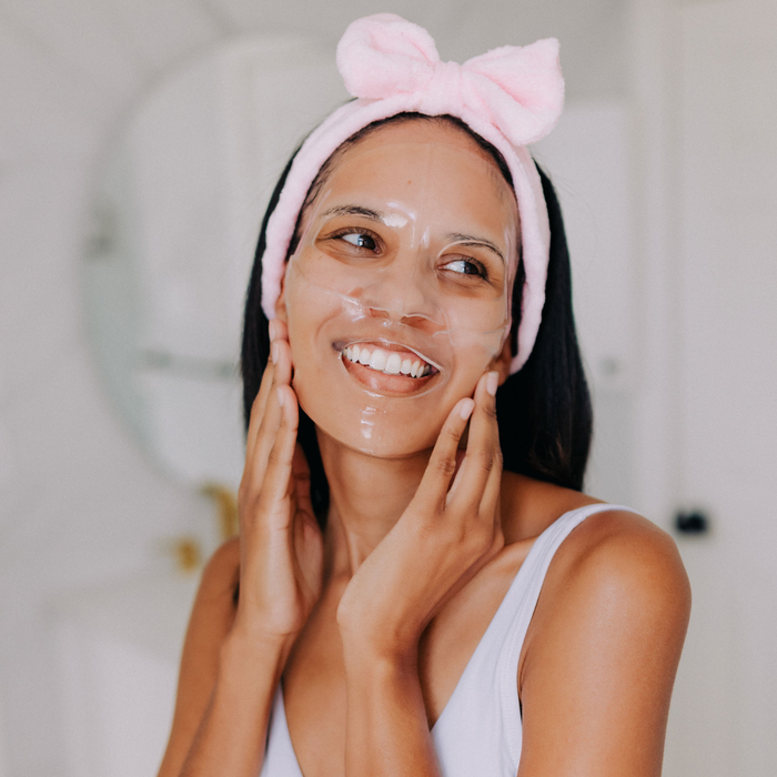 Woman applying a full face collagen mask to her face with a pink headband, smiling in front of a mirror.