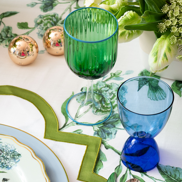 Green glass goblet with vertical ridges next to a round blue glass goblet with a thick base on a floral tablecloth.