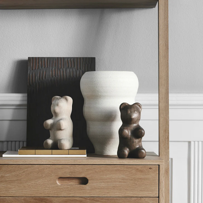 Interior shelf arrangement featuring two small bear-shaped wooden figurines in natural oak and dark-toned wood, placed beside a sculptural white ceramic vase and a textured dark panel, styled on stacked books within a light wood shelving unit against a soft gray wall.