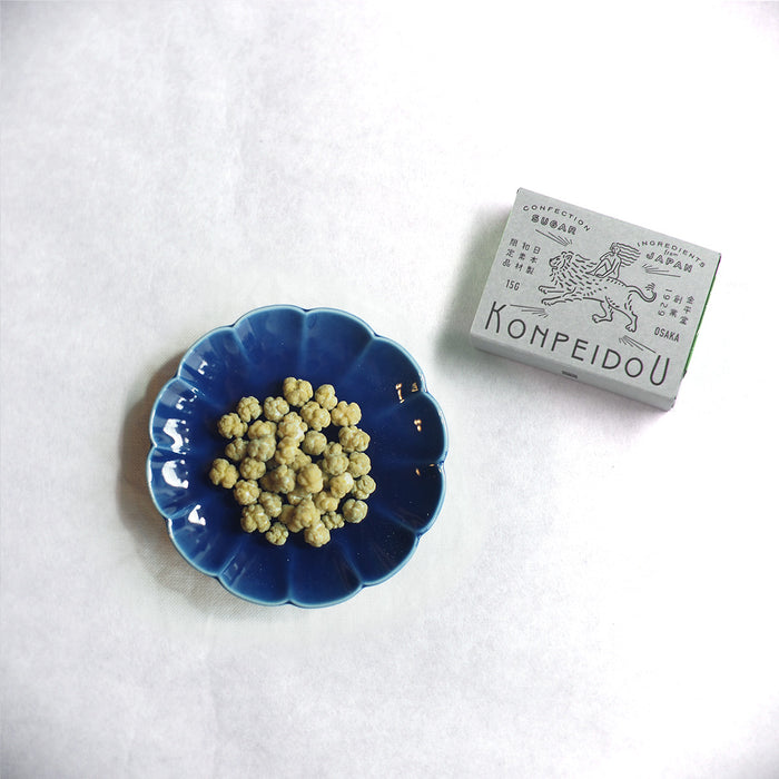 A small blue scalloped dish filled with round pale yellow beads or seeds, placed on a white textured surface next to a gray box labeled KOMPEIDOU.