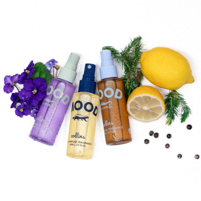 Three Mood Water all-purpose fragrance sprays arranged with violet flowers, lemon, juniper berries, and evergreen sprigs, fresh botanical product flat lay on a clean white background.