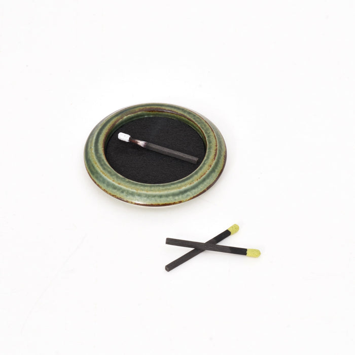 A green ceramic dish on a white background holding a round black striker pad with one incense match resting inside, accompanied by two loose incense matches placed nearby.