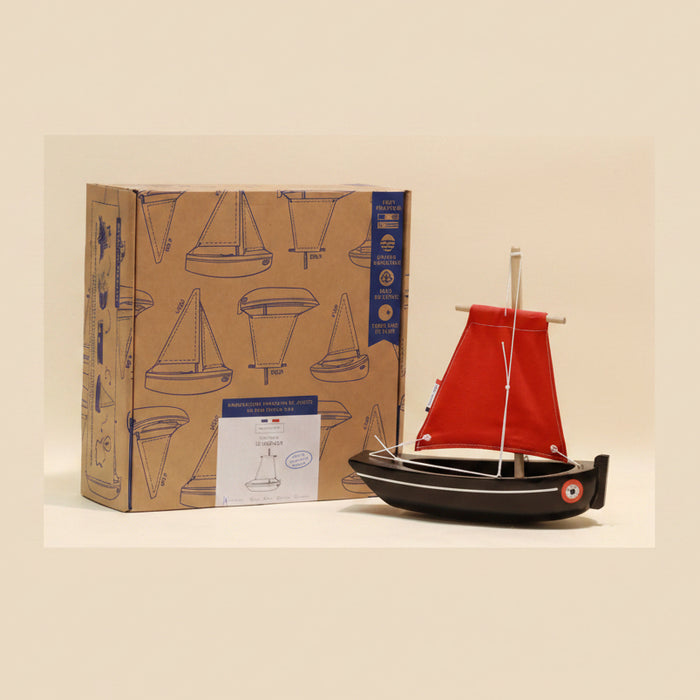 Model sailboat with red sail next to its packaging box on a beige background