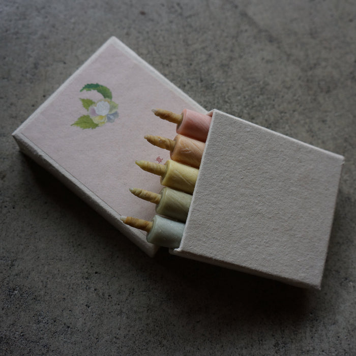 A matchbox-style candle holder with five wax candles inserted, photographed on a textured gray surface. The box is off-white with a small green and blue floral illustration on the lid, and the visible wax colors include pink, yellow, cream, and pale green.