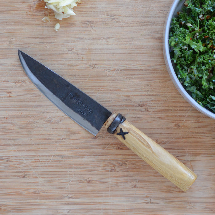 Small hand-forged kitchen knife with a dark steel blade and wooden handle, displayed on a cutting board beside chopped garlic and salad in a rustic kitchen setting.