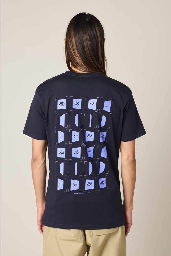 A person with long hair, seen from behind, wears a navy blue 686 24/7 Authentic SS Tee by 686, made from 100% cotton and featuring blue and purple geometric patterns on the back, paired with beige pants against a plain light background.