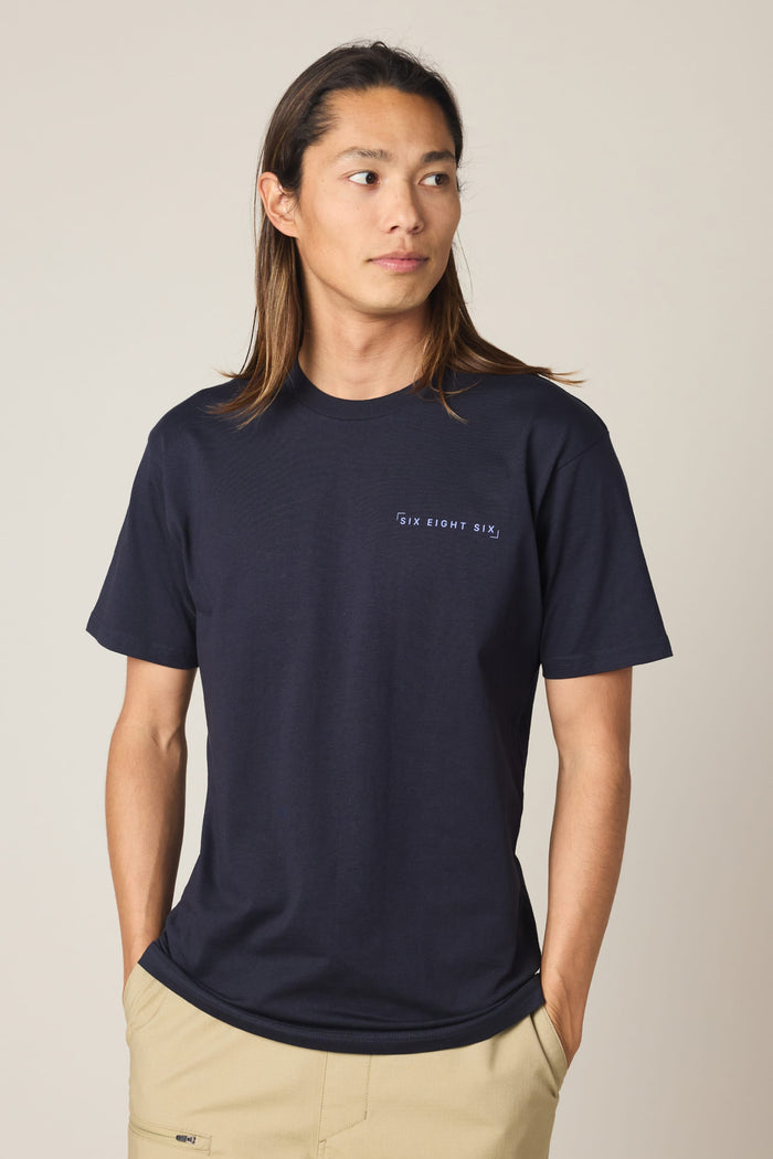 A person with long brown hair wears the 686 24/7 Authentic SS Tee by 686‚Äîa navy blue men‚Äôs t-shirt with ‚ÄúSIX EIGHT SIX‚Äù on the chest‚Äîpaired with beige pants, hands in pockets, looking sideways against a plain background. Made from 100% cotton.