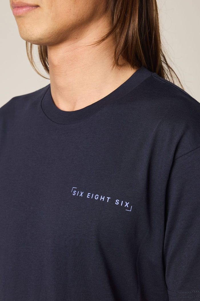A person with long brown hair wears a 686 24/7 Authentic SS Tee‚Äîa dark navy men's t-shirt made from 100% cotton with "SIX EIGHT SIX" in white on the chest. The image shows the upper torso and part of their face.