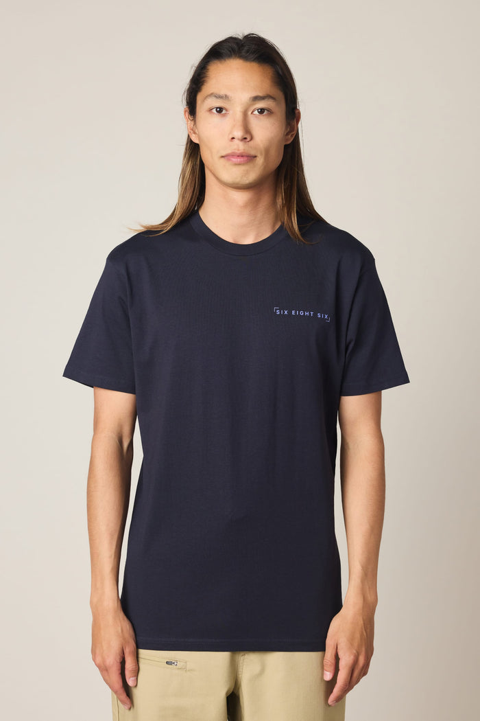 A person with long brown hair stands facing forward against a plain light background, wearing the 686 24/7 Authentic SS Tee by 686‚Äîa navy blue men's t-shirt made from 100% cotton with small white text on the left chest‚Äîand beige pants.