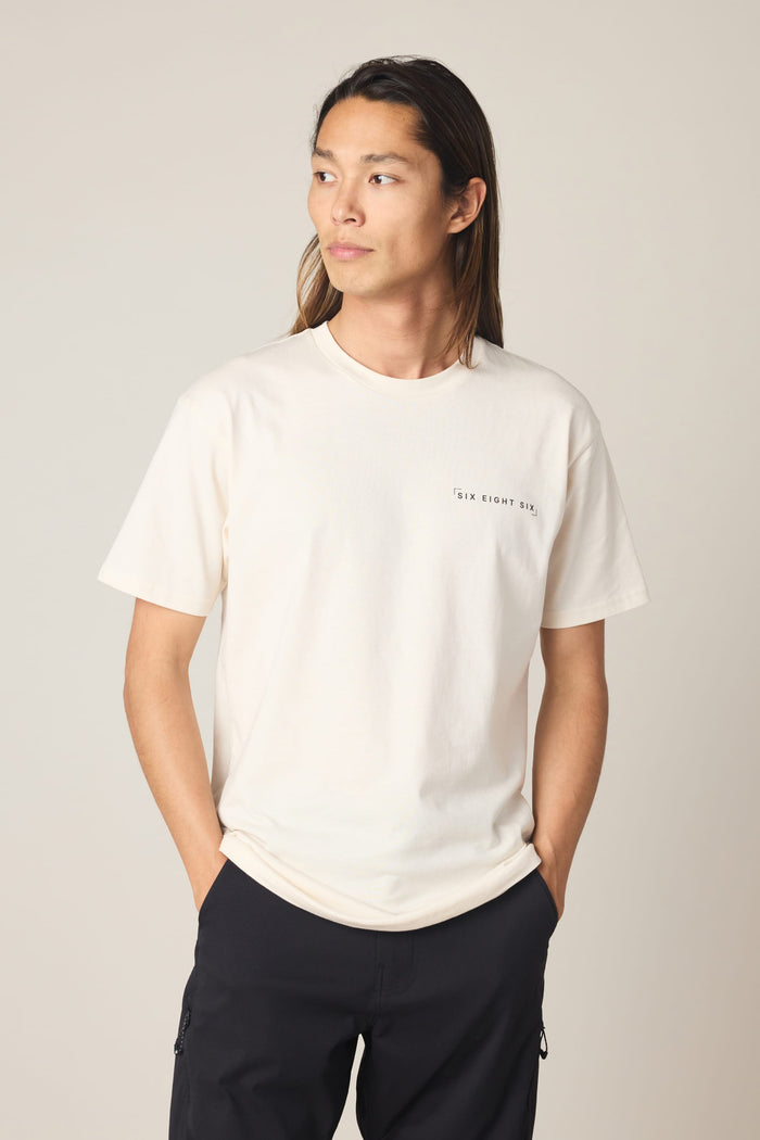 A person with long hair stands against a plain background, hands in pockets, wearing the 686 24/7 Authentic SS Tee by 686‚Äîa cream men's cotton t-shirt with small chest text‚Äîpaired with dark pants, looking slightly to the side.
