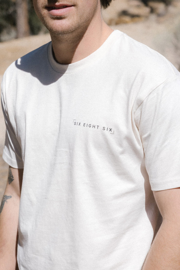 A person wearing the 686 24/7 Authentic SS Tee by 686, a white 100% cotton men's t-shirt with "SIX EIGHT SIX" in small black letters on the front, is seen outdoors in natural light. Only their upper body is visible.