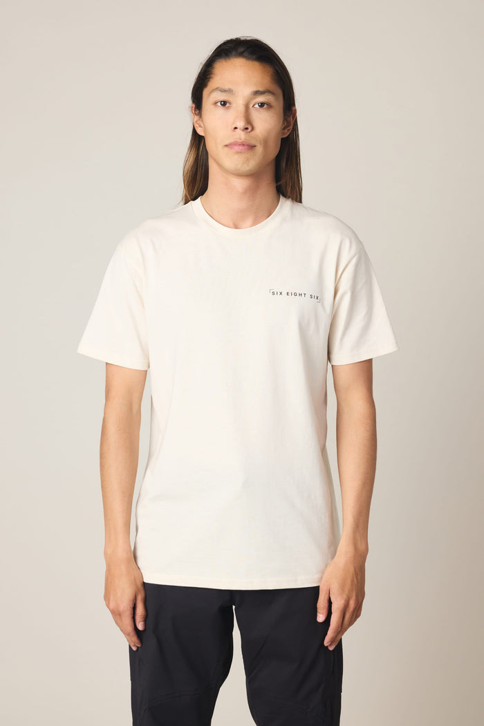 A person with long hair faces forward, wearing the 686 24/7 Authentic SS Tee by 686‚Äîan off-white men's t-shirt‚Äîpaired with black pants, against a simple light gray background.