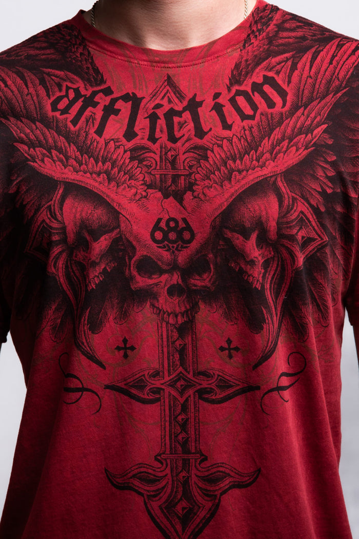 A person wearing the 686 Affliction Death Eyes SS Tee by 686, a men‚Äôs 100% cotton shirt with black gothic artwork‚Äîskulls, wings, crosses, and "Affliction" across the chest. Image shows only the chest and neck.