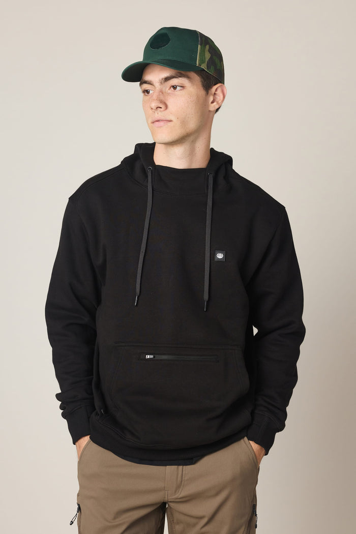 A young man stands with his hands in his pockets, looking to the side, wearing the 686 Men's All Season Hoody in black and a green camo cap. He pairs the hoodie with tan pants and poses against a plain, light-colored background.