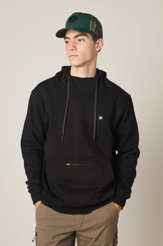 A young man stands with his hands in his pockets, looking to the side, wearing the 686 Men's All Season Hoody in black and a green camo cap. He pairs the hoodie with tan pants and poses against a plain, light-colored background.