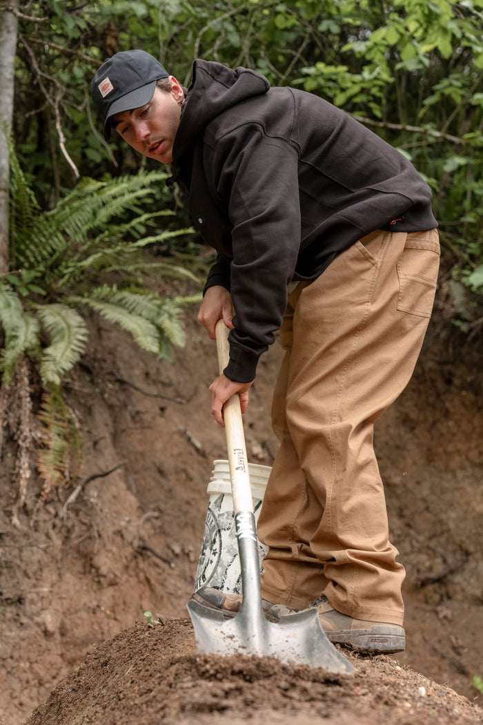 A person wearing the 686 Men's All Season Hoody by 686, along with tan pants and a black cap, is standing outdoors, leaning on a shovel and digging in the dirt near green foliage and a forested backdrop.
