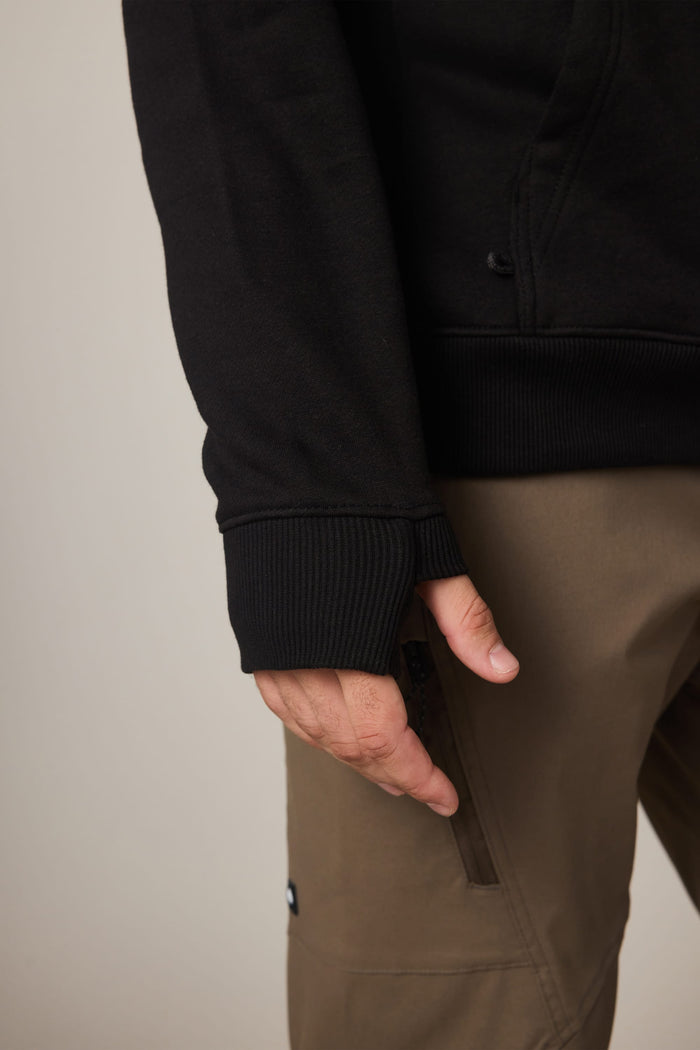 A person wearing the 686 Men's All Season Hoody in black with thumbholes and brown pants, shown from the waist down with their left hand relaxed by their side.