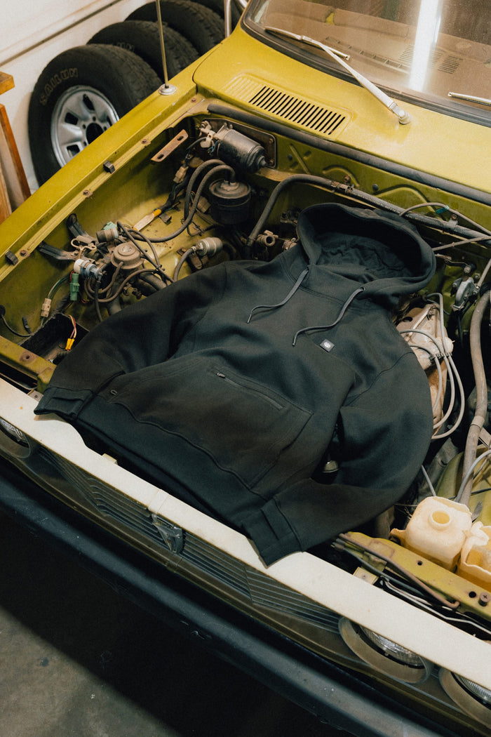 A 686 Men's All Season Hoody in black is laid out over the engine bay of a yellow car with its hood open inside a garage.