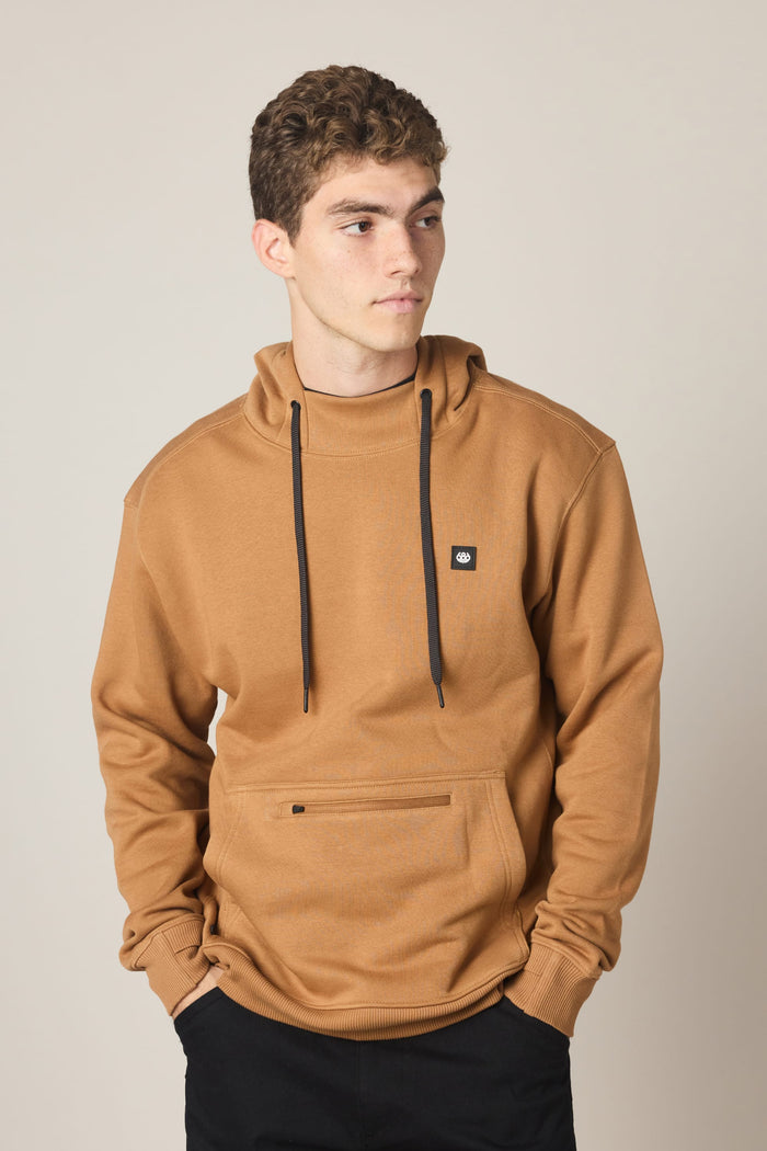 A young man with curly brown hair wears the 686 Men's All Season Hoody—a tan sweatshirt by 686 featuring black drawstrings and a front pocket. He stands with his hands in the pockets, looking to his left against a plain background.