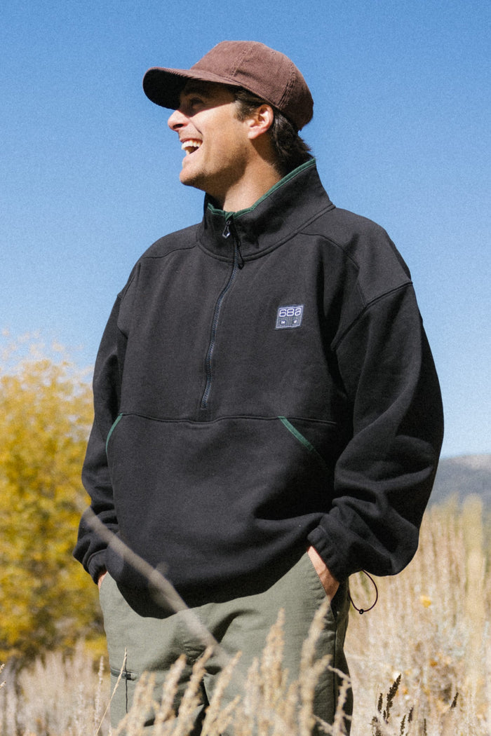 A man wearing the 686 Men's Burnout Anorak stands outdoors, smiling and looking to the side. Tall grass and colorful trees are visible under a clear blue sky, providing the perfect backdrop for this stylish jacket by 686.