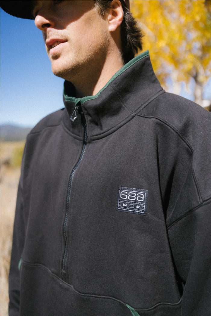 A man stands outdoors wearing the 686 Men's Burnout Anorak, a black zip-up jacket from 686 that features a waterproof design reminiscent of classic men's outerwear. The scene is set against yellow autumn leaves and a clear blue sky, with only the lower half of his face visible.