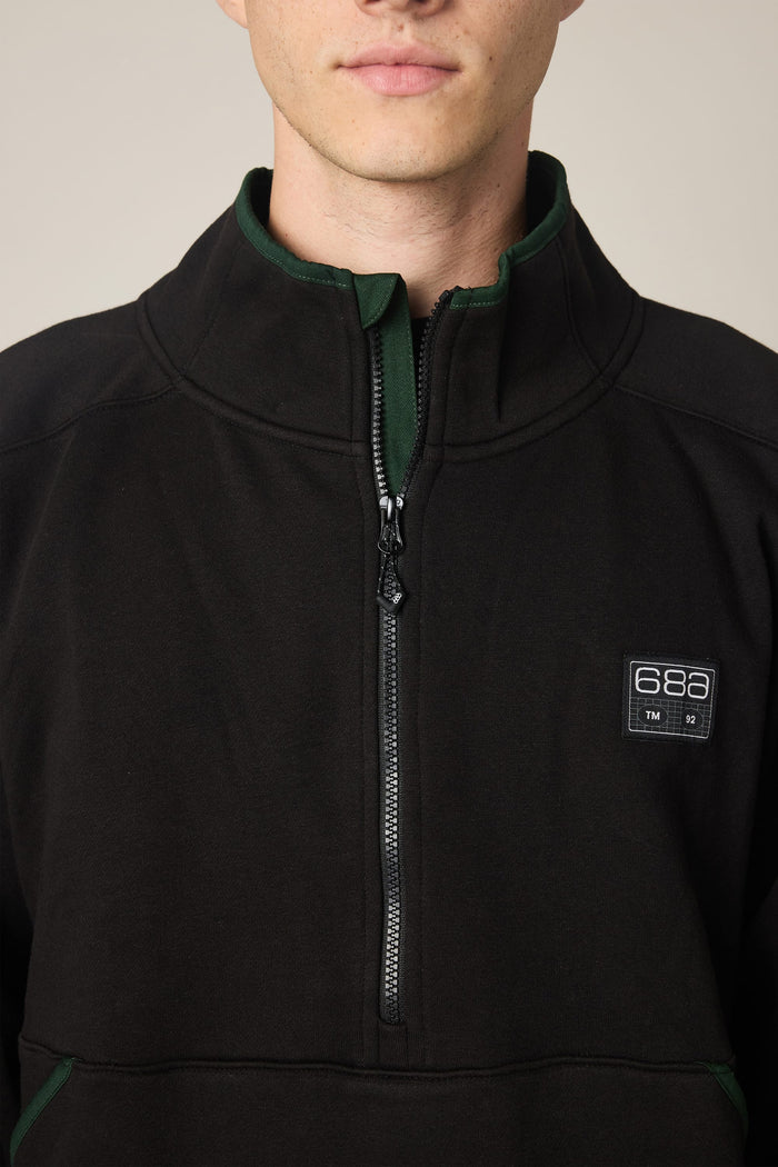 A man wearing the 686 Men's Burnout Anorak—a black half-zip sweatshirt with green trim and a 686 logo patch on the chest—layered over a men's woven jacket. Only the lower half of his face and upper torso are visible.