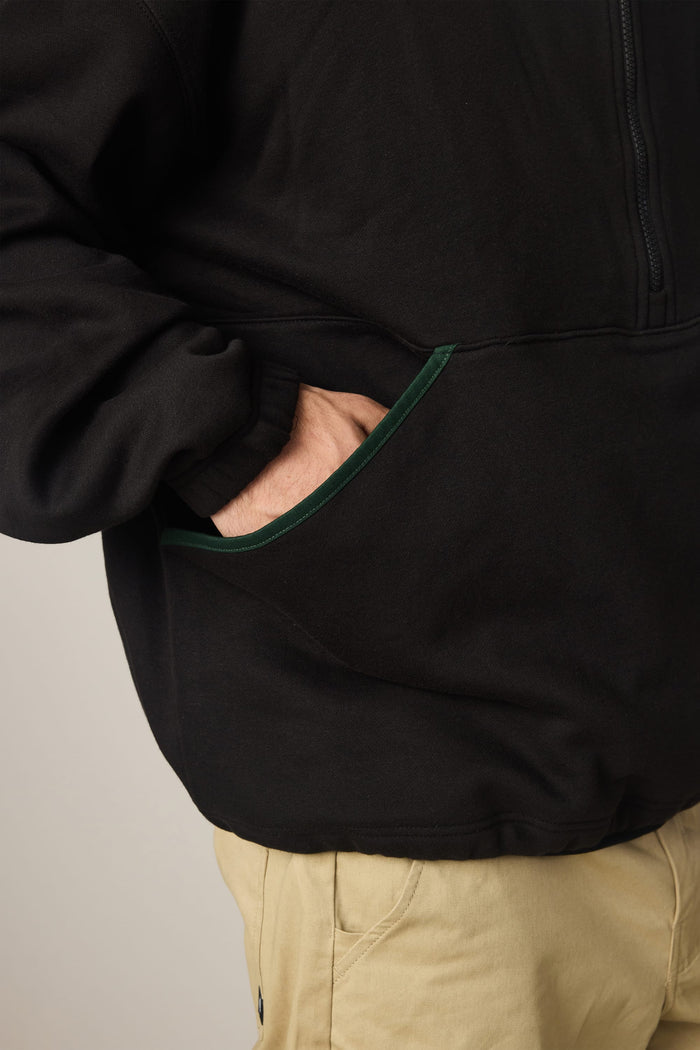 A person wearing the 686 Men's Burnout Anorak—an all-black waterproof jacket by 686—with one hand in a side pocket, paired with light tan pants. Only the torso and upper legs are visible.