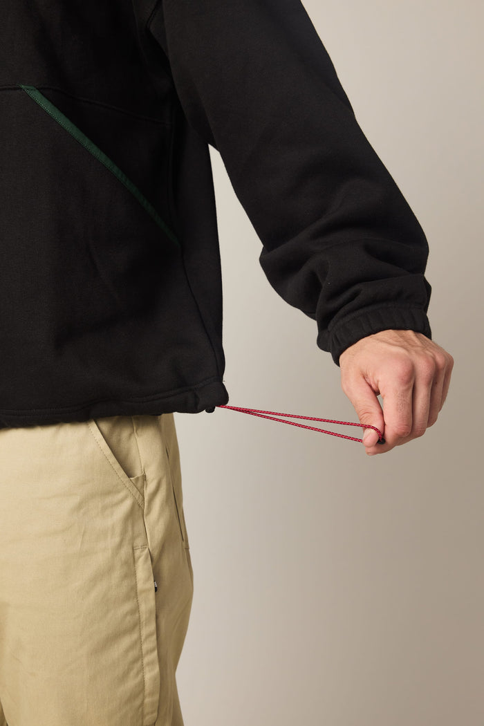 A person wearing the 686 Men's Burnout Anorak in black and beige pants adjusts the red drawstring at the jacket's hem, tightening it with one hand.