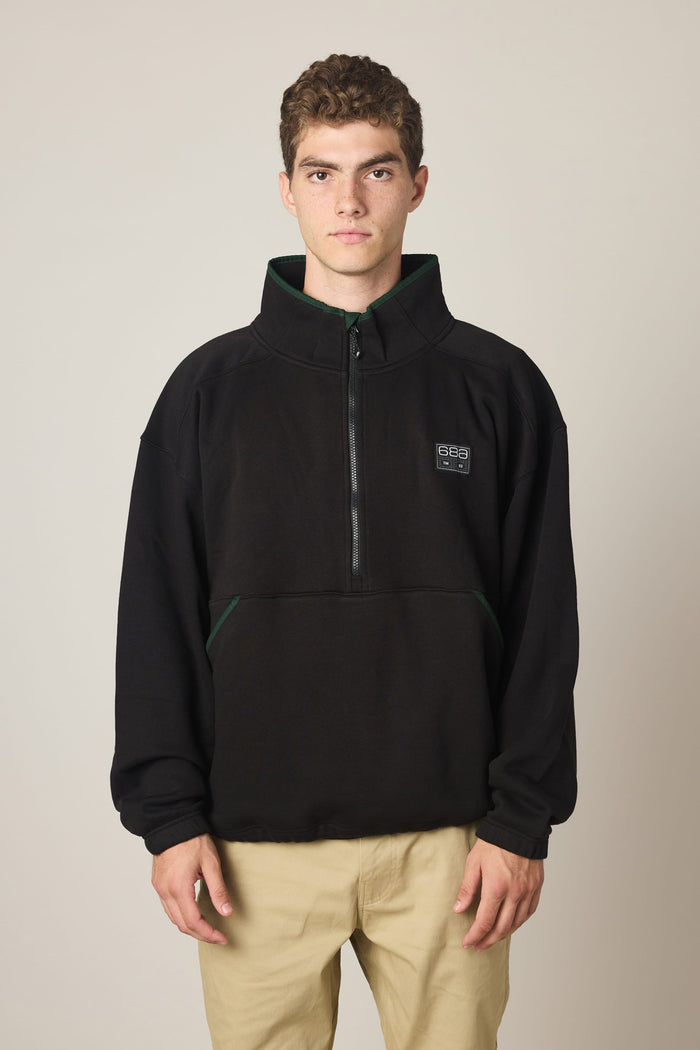 A young person with short curly hair wears the 686 Men's Burnout Anorak—a black waterproof jacket from 686 featuring a small logo—paired with beige pants, standing against a plain light background and looking directly at the camera with a neutral expression.