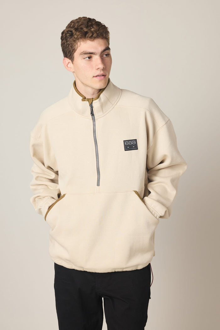 A young man stands with his hands in the pockets of the 686 Men's Burnout Anorak, a light beige woven jacket from 686 featuring a quarter-zip pullover design with a high collar and front pocket, looking to his left against a plain background.