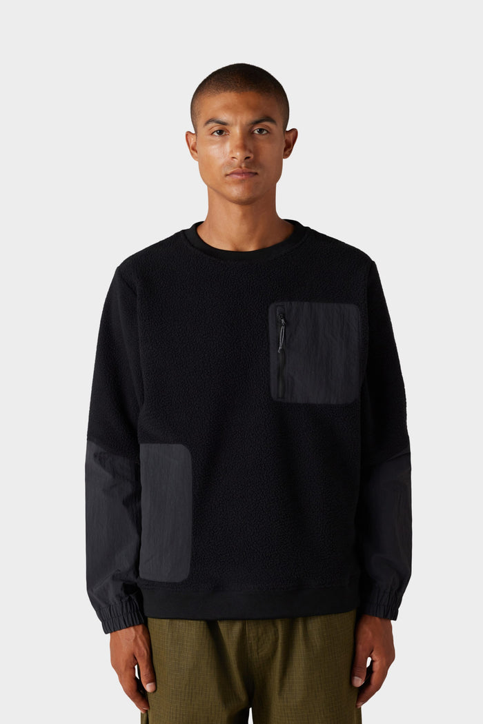 A person wearing the 686 Mens Buttermilk Sherpa Crew‚Äîa black textured long-sleeve sweatshirt‚Äîpaired with olive-green pants, stands against a plain light gray background.