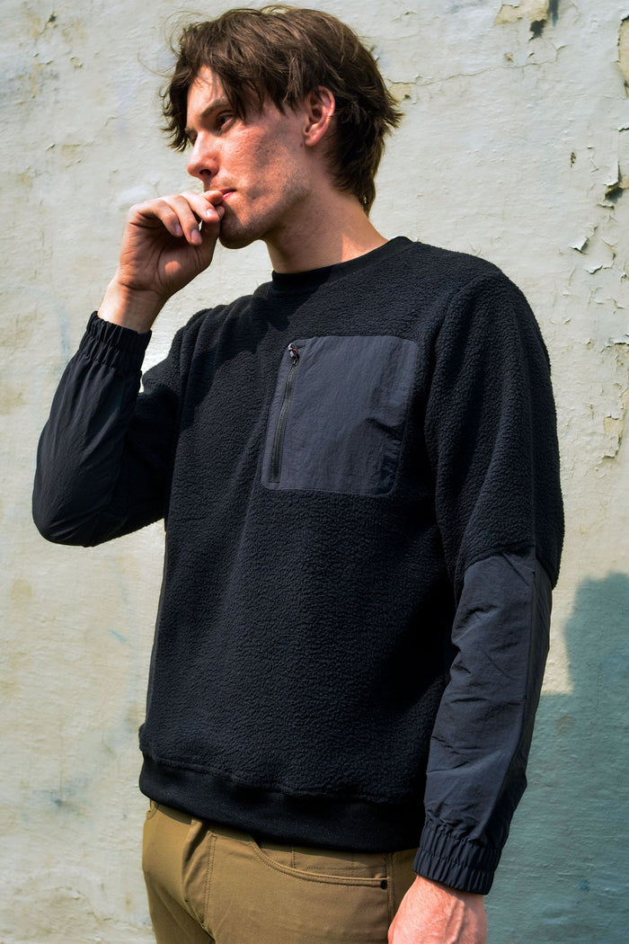 A man with tousled hair stands against a light, weathered wall in khaki pants, wearing the 686 Mens Buttermilk Sherpa Crew for style and comfort, holding his hand near his mouth as if smoking.