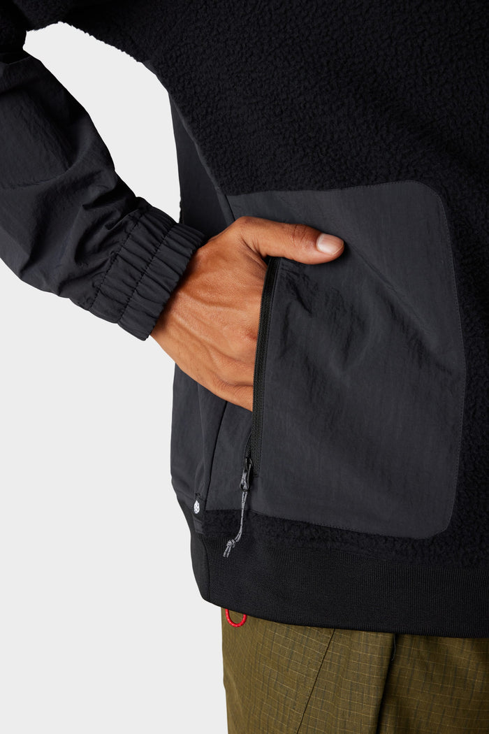 A person zips the pocket of a 686 Mens Buttermilk Sherpa Crew, featuring a textured pocket and elastic cuff, while wearing olive green pants.