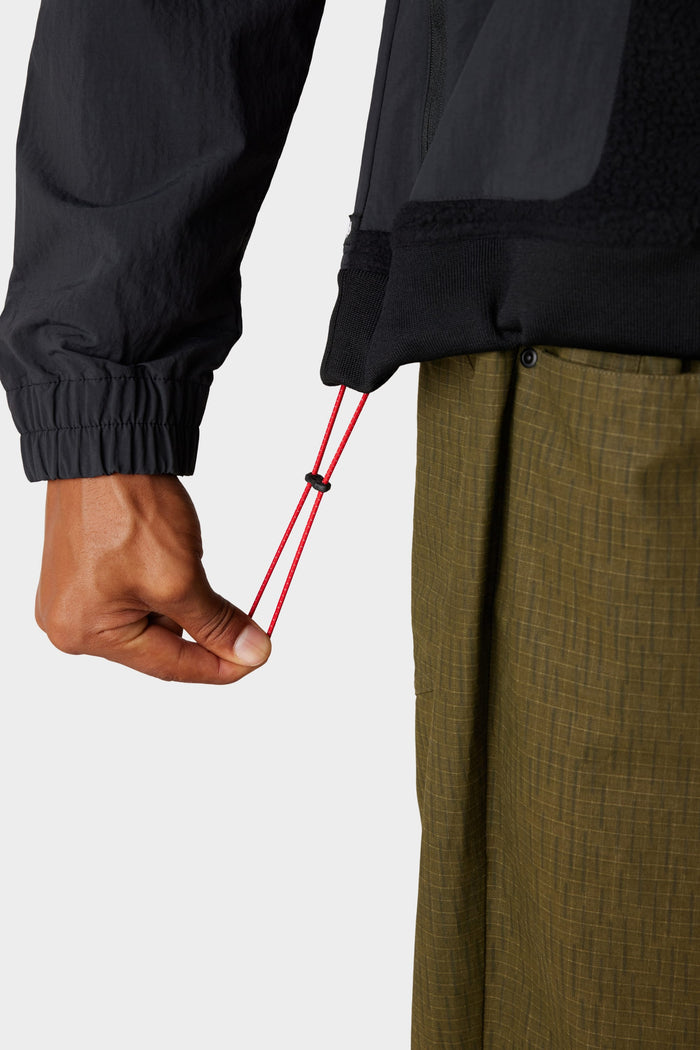 A person in the 686 Mens Buttermilk Sherpa Crew in olive-green adjusts the red drawcord on a black jacket hem, with only their hand, sleeve, and pants visible‚Äîshowcasing comfort and style from 686.