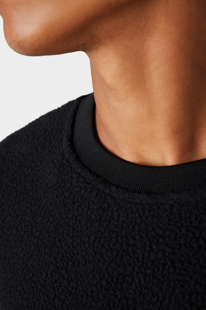 A close-up of a person‚Äôs neck and lower face wearing the 686 Men‚Äôs Buttermilk Sherpa Crew, a textured black sweater by 686, that combines comfort and style against a plain light background.