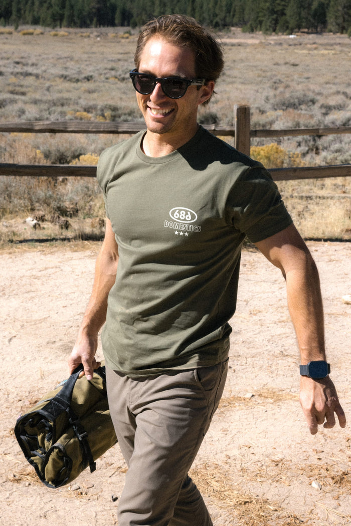 A smiling man wearing sunglasses and the 686 Men's Domestics Authentic SS Tee, a 100% cotton green t-shirt by 686, along with khaki pants, walks outdoors on a sunny day holding a rolled-up green bag. A wooden fence and trees can be seen in the background.