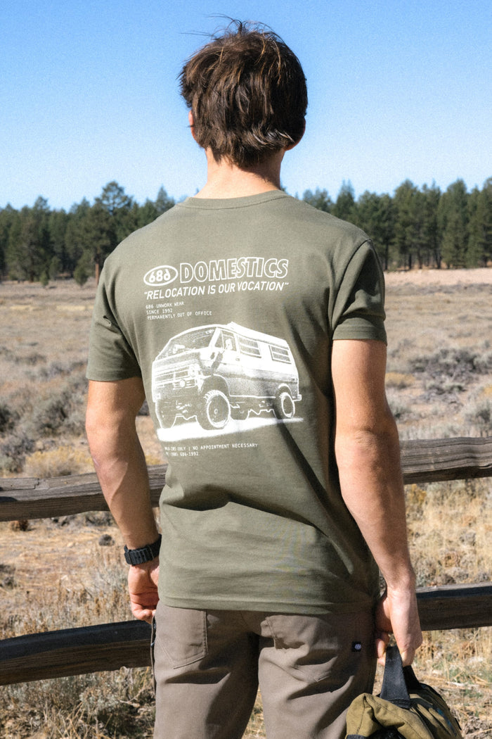 A person stands outdoors by a wooden fence, facing away, wearing the 686 Men's Domestics Authentic SS Tee. This olive-green 100% cotton t-shirt from 686 features a graphic of a camper van with the words “688 DOMESTICS ‘RELOCATION IS OUR VOCATION,’” set against trees and fields in the background, offering comfort and style for any adventure.