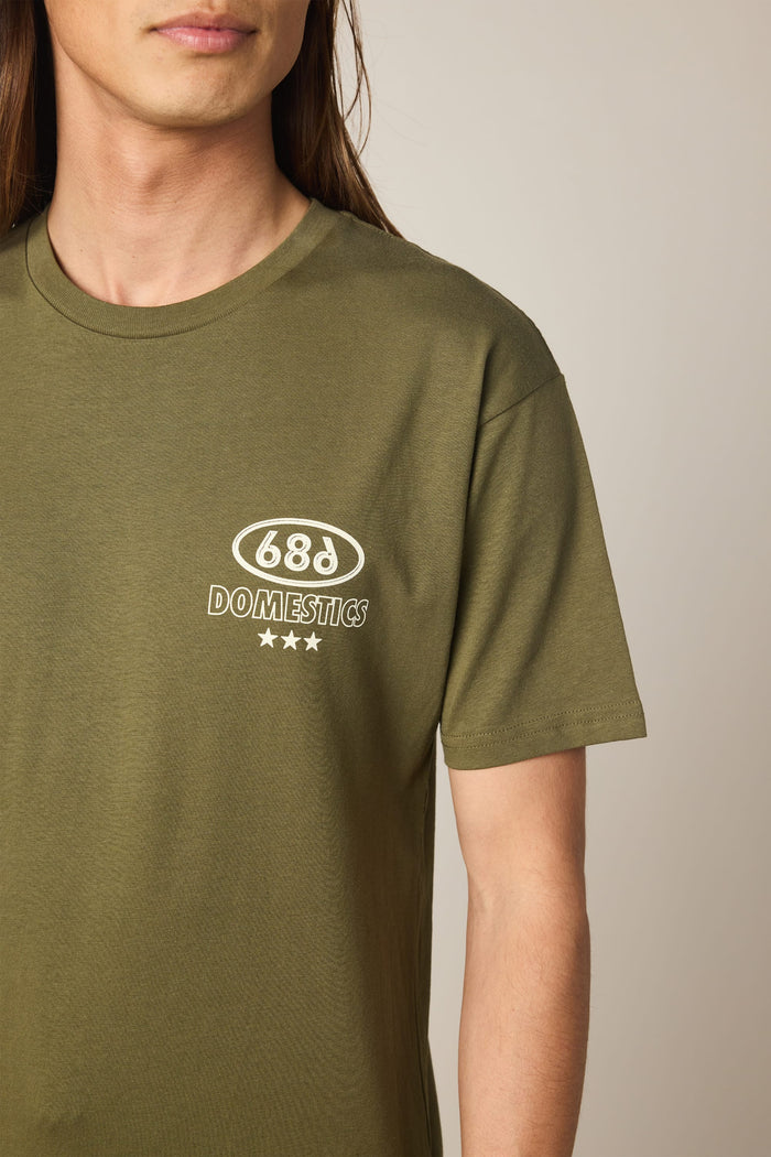 A person with long hair wears the 686 Men's Domestics Authentic SS Tee, an olive green cotton t-shirt featuring the 686 DOMESTICS logo and three white stars on the left chest. Made from 100% cotton, this shirt by 686 offers both comfort and style.