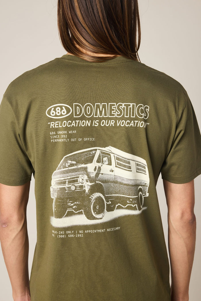 A person is shown from the back wearing the 686 Men's Domestics Authentic SS Tee by 686—an olive green, 100% cotton T-shirt featuring a graphic van and the text “DOMESTICS” and “RELOCATION IS OUR VOCATION,” along with additional smaller brand details.