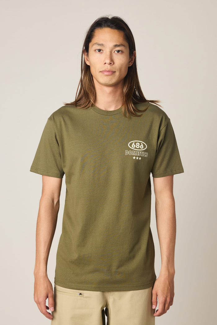 A person with long hair wears the 686 Men's Domestics Authentic SS Tee—an olive green 100% cotton T-shirt by 686 featuring a small white logo and text on the left chest—while standing against a plain light background.