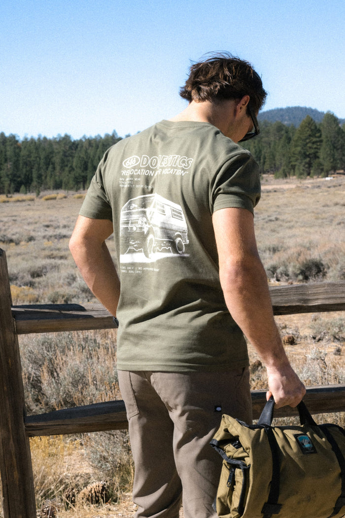 A person stands outdoors by a wooden fence, facing away, wearing the 686 Men's Domestics Authentic SS Tee in olive green and holding a green bag, with trees and mountains visible in the background.