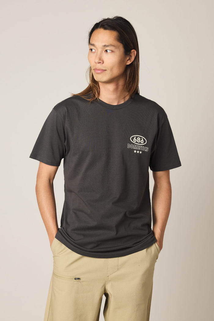 A person with long hair stands with hands in their pockets and looks to the side against a plain background, wearing tan pants and a black 686 Men's Domestics Authentic SS Tee by 686.