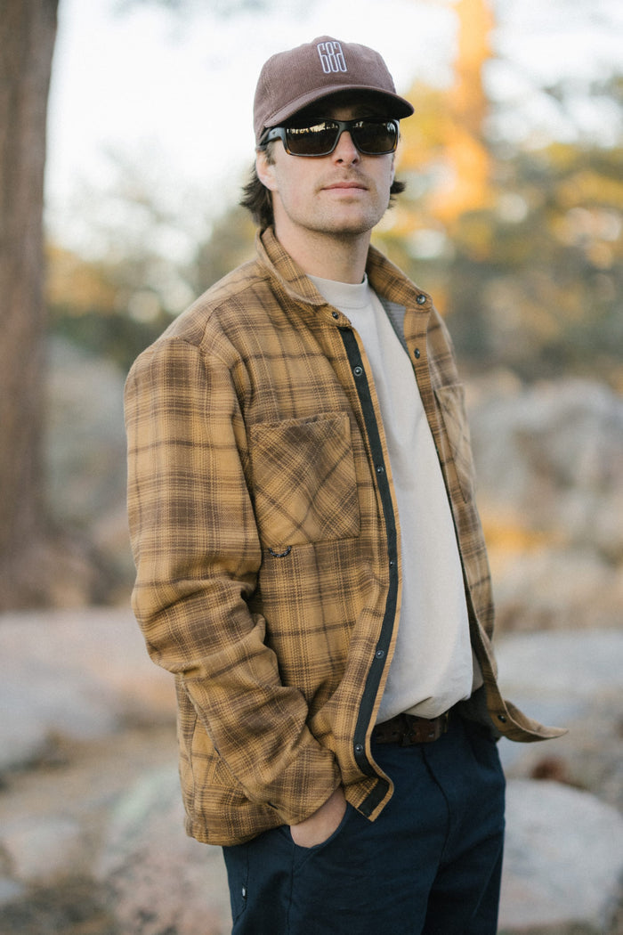 A man wearing sunglasses, a brown plaid jacket, the 686 Men's Heat Grid™ Flannel—a light and comfortable polyester knit shirt by 686—and a brown cap stands outdoors with his hand in his pocket. Trees and rocks are blurred in the background.