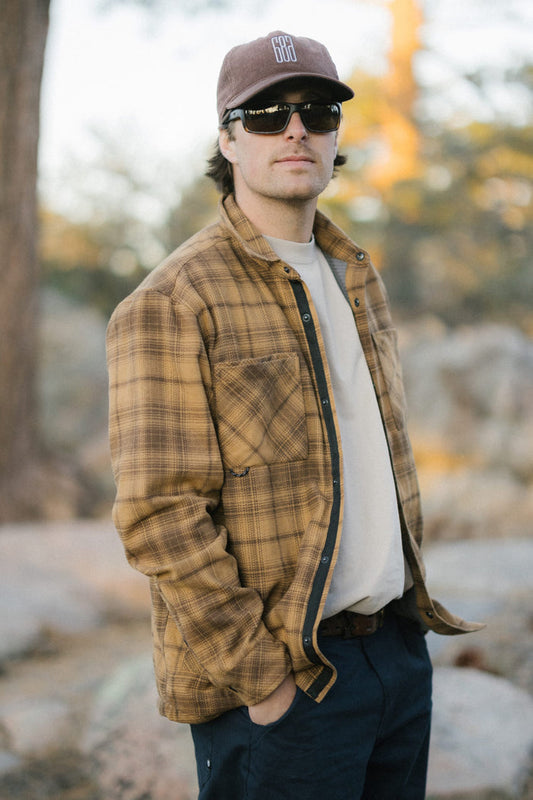 A man wearing sunglasses, a brown plaid jacket, the 686 Men's Heat Grid™ Flannel—a light and comfortable polyester knit shirt by 686—and a brown cap stands outdoors with his hand in his pocket. Trees and rocks are blurred in the background.