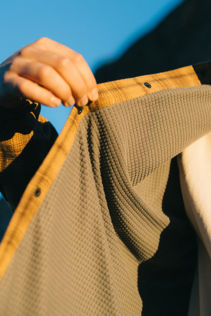 A hand holds open a jacket, revealing its textured Heat Grid™ waffle-patterned gray lining and soft inner layer—evoking the comfortable finish of a 100% polyester shirt from 686's Men's Heat Grid™ Flannel—while sunlight highlights the fabric details.