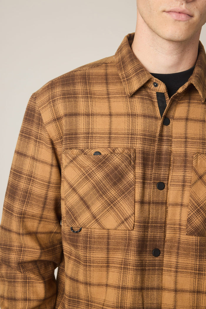 A person wearing the 686 Men's Heat Grid™ Flannel, a brown plaid button-up shirt from 686 featuring two chest pockets, is shown from the shoulders to mid-torso with part of the face visible. This stylish look offers comfort and durability thanks to its polyester knitted construction.
